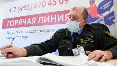 En säkerhetsvakt i uniform och munskydd antecknar i en bok. Han sitter framför ett plakat med text på ryska.