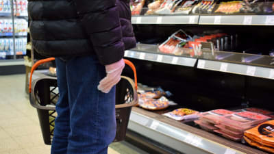 En äldre person står vid köttdisken i matbutiken med plasthandske på handen.