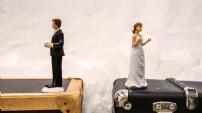 Ett bröllopspar (två figurer, kakdekorationer) står på kappsäckar och tittar bort från varandra.