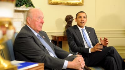 Bild på två män i kostym och blå slipsar som sitter bredvid varandra och diskuterar.