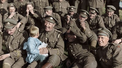 Soldater sitter uppställda och spelar munspel och skrattar, en av soldaterna håller ett litet barn i famnen.