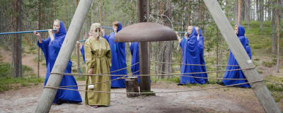 Metsässä sinisiin kaapuihin pukeutuneita naisia, soittavat sinisiä huiluja, keskellä ison kirkonkellon äärellä vihreäkaapuinen nainen naamio kasvoilla. Soivan Metsän Ilmattaria.