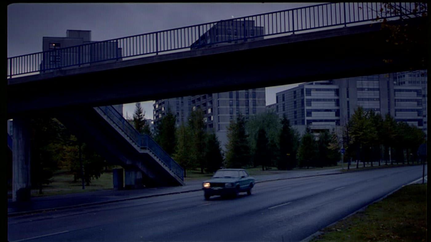 Sininen Ford Taunus, jonka toinen etuvalo on sammunut, ajaa yksinäisellä, aamuöisellä Sörnäisten rantatiellä Helsingissä, jalankulkijasillan alla, taustalla kohoaa Merihaka.