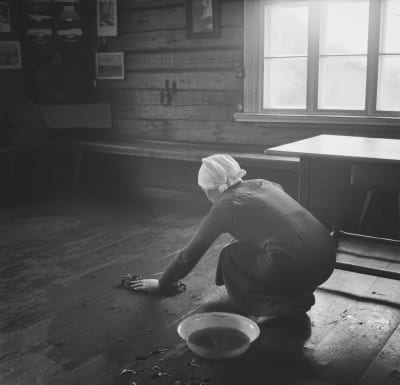 Svartvit bild på kvinna som skurar golvet i ett torp på 1930-talet.
