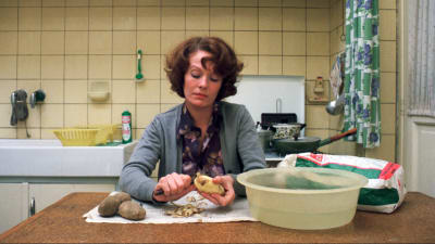 Näyttelijä Delphine Seyrig istuu keittiön pöydän ääressä ja kuorii perunoita. Kuva elokuvasta Jeanne Dielman.