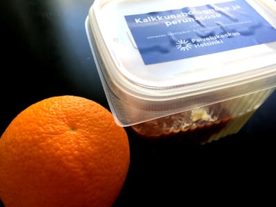 En förpackning med skolmat. Bredvid plastlådan sitter en apelsin.