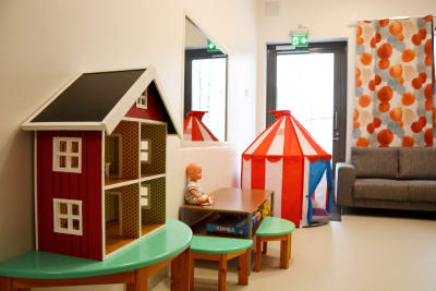 ett rum med ett dockhus, en docka, en spegel, ett litet cirkustält, en soffa och gardiner med orangeröda detaljer på