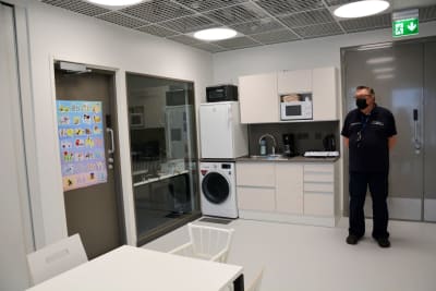 ett person står i ett kök med köksutrustning och tvättmaskin, bord och stolar och en dörr till ett övervakningskontor i ett fängelse