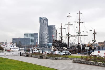 Det svarta piratskeppet Dragon ligger vid kajen i Gdynia, med gräsmatta och bilar framför och höghus i bakgrunden.