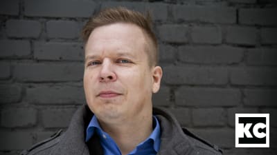 Kirjailija Ville Hytönen seisoo harmaan tiiliseinän edessä ja katsoo kameraan.