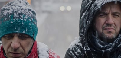 Kaksi miestä lähikuvassa lumisateessa, toinen katsoo alas, toinen hupun alta kameraan.