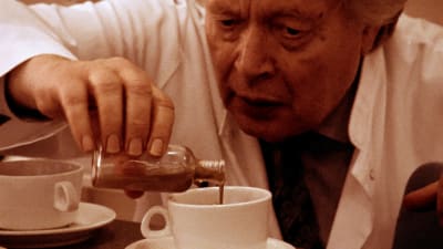 Närbild på Stig Helmer (Ernst Hugo Järegård) som häller någon konstig vätska ur en glasflaska i en kaffekopp.