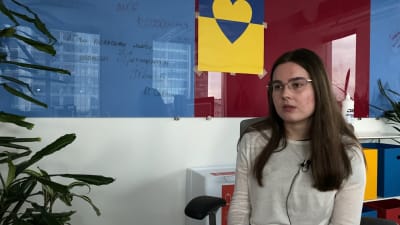 En kvinna med långt hår, vit tröja och glasögon talar. Hon sitter inomhus, i väggen bakom finns en bild på Ukrainas flagga och ett hjärta i blått och gult.