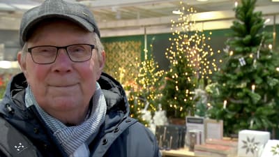 En äldre man med glasögon och keps. I bakgrunden julgranar i ett köpcentrum. 