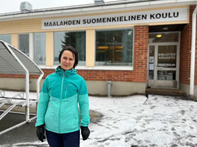 Kvinna står utomhus framför en röd tegelbyggnad med texten Maalahden suomenkielinen koulu.