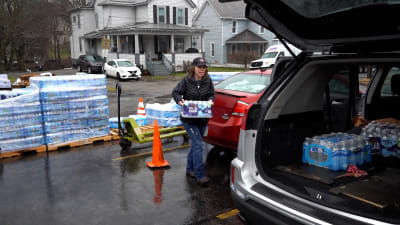 En kvinna bär en back med vattenflaskor till sin bil från en rad lastpallar med vattenbackar. I den öppna bakluckan syns två backar till.