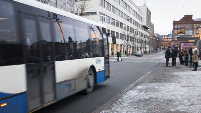 En buss kör mot en busshållplats där personer står och väntar.