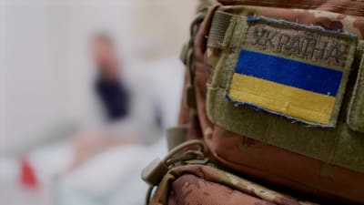 Nuhjuiseen armeijareppuun kiinnitetty ukrainan lippu tai Ukrainan armeijan tunnus. Sumeasta taustasta saattaa erottaa istuvan henkilön.