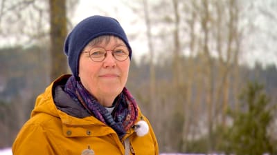 En kvinna i blå mössa och gul jacka tittar in i kameran. 