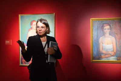 En medelålders, glasögonförsedd kvinna i ett museum med en bok under sin vänstra arm medan hon gestikulerar med sin högra arm och ser ut att kommunicera med museibesökare. 