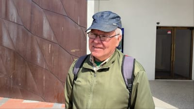 En äldre man i keps och glasögon står framför en modern byggnad. 