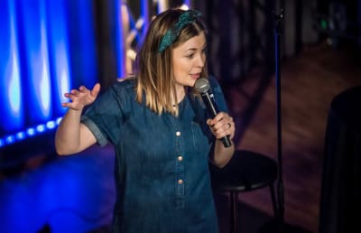 Anna Rimpelä står på en scen med mikrofonen i handen. Hon gestikulerar med högra handen och i bakgrunden syns ett blått draperi.