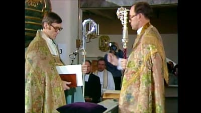 John Vikström viger Erik Vikström till biskop i en kyrka. Båda har broderade kåpor på sig och en biskopsstav i handen.