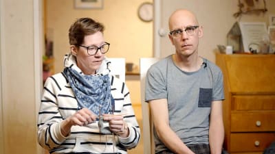 Heidi och Andreas Forsman sitter tillsammans. Heidi i randig tröja och glasögon sitter och stickar, Andreas i grå t-skjorta och glasögon ser mot kameran. Båda ser allvarliga ut.