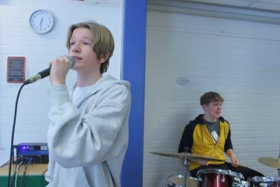 Två ungdomar, pojken längst fram står med mikrofon och sjunger och pojken bakom sitter och spelar trummor.