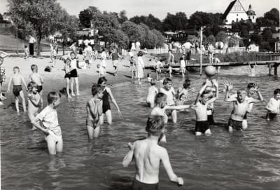 Svart-vit bild av barn och unga som leker i vattnet vid en badstrand på 1960-talet.