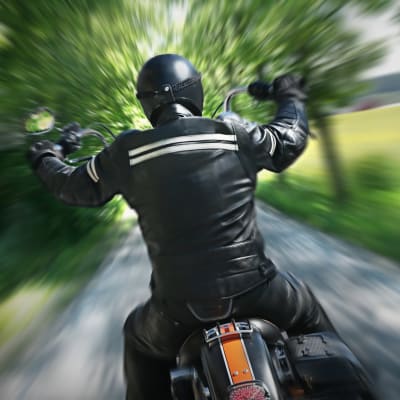 En motorcyklist i svart hjälm och svarta läderkläder sedd bakifrån, kör på en smal väg med träd och grönska på båda sidorna. Skakig bild.