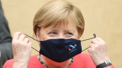 Merkel riisuu maskia, jossa on Saksan EU-puheenjohtajuuslogo.