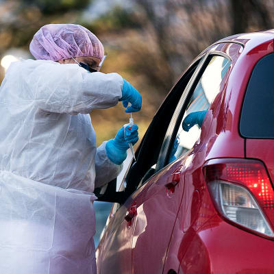 En person i skyddskläder tar ett coronavirusprov med en bomullspinne av en person i en bil med nedvevad bilruta.