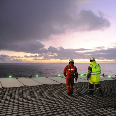 Två personer, den ena i röd overall och den andra i gul, går på däcket till en oljeplattform i solnedgången över Atlanten.