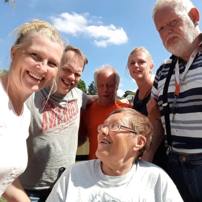 Medelålders och lite äldre personer med funktionsnedsättning samt två yngre vårdare ser glada ut i en gruppbild. Sol och sommar.