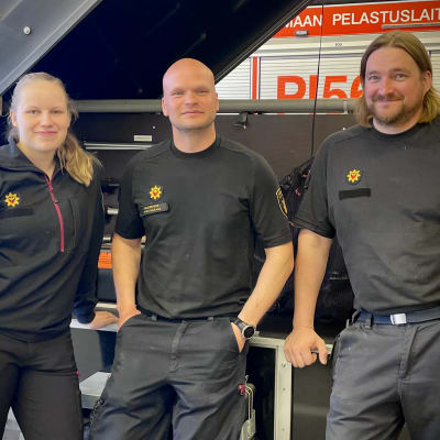 Kolme pelastajaa nojaa kuljetuskärryyn ja katsoo kameraan