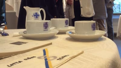 Kaffekoppar och en kaffekanna samt en servett med Lotta Svärd-emblem. Äldre kvinnor syns i bakgrunden.