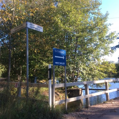 En bro leder över en sjö, gatunamnsskyltar, en skylt som visar vägen till Katinsuo (Kattkärret), trafikmärke för återvändsgränd, soligt höstväder