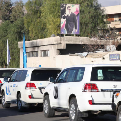 YK:n henkilöautoja kulkee ohi rakennuksen, jota koristaa presidentti Bashar al-Assadin kuva.