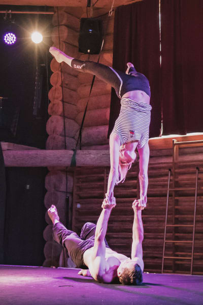 Kaksi sirkustaiteilijaa tekemässä temppua, nainen seisoo käsillä selällään maassa makaavan miehen käsien varassa