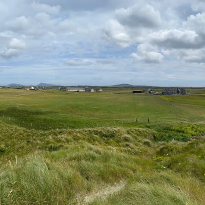 Maisemakuva Ulko-Hebrideiltä. Kuvassa vihreä niitty, asutusta kaukana ja pilviä taivaalla.