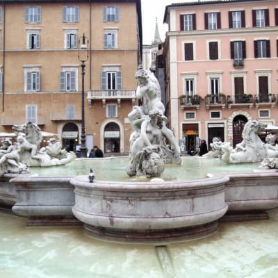 Monin veistoksin koristeltu suihkulähde Piazza Navonalla Roomassa, taustalla punertavia rakennuksia.