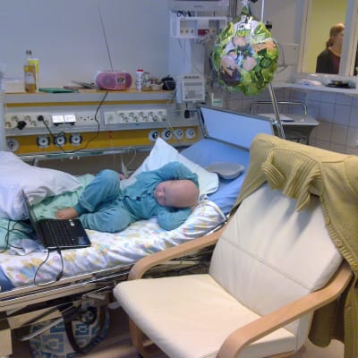 Cancersjukt ban ligger i patientsäng på sjukhus och använder bärbar dator.