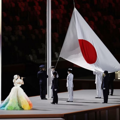 Den japanska nationalsången sjungs av en en kvinna i fluffig och färggrann klänning när den japanska flaggan hissas inne på olympiastadion i Tokyo.