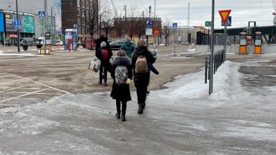En grupp med personer, en familj, med ryggarna mot kameran, går bort. Det är i Helsingfors. Alla bär en ryggsäck eller väska..