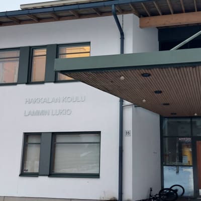 Koulun vaalea seinä ikkunoineen, ovi sekä oven päällä katos ja seinässä teksti Hakkalan koulu, Lammin lukio.