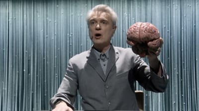 Harmaapukuinen David Byrne laulaa puolikuvassa lavalla aivot kädessä