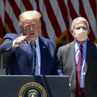 Donald Trump står vid ett talarpodium och håller presskonferens. I bakgrunden syns smittskyddsexperten Anthony Fauci som bär ansiktsskydd. Trump är utan.
