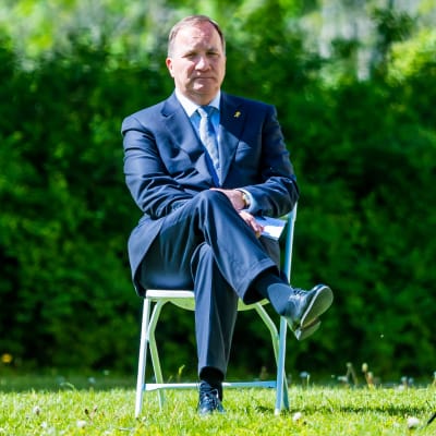 Stefan Löfven sitter på en stol på en gräsmatta. Han sitter med benen i kors och är klädd i kostym.
