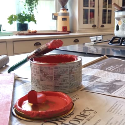 En pensel som doppats i i rosa färg är ställd mot en öppnad burk med målarfärg. Målarfärgsburken står på en köksbänk som är täckt med tidningspapper. I bakgrunden står en kastrul på spisen.